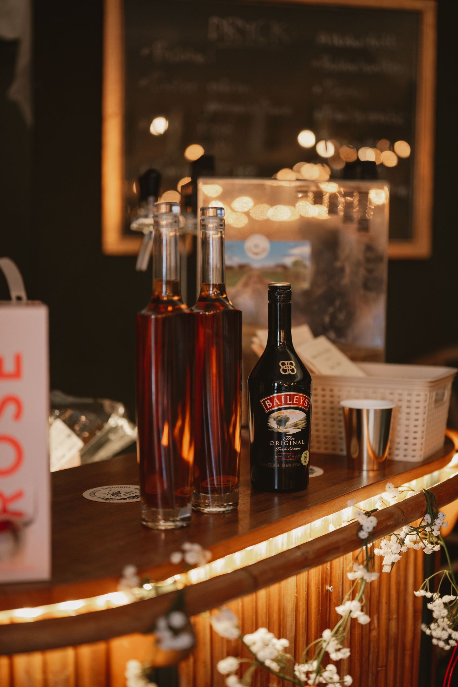Baileys Irish Cream bottle on a warmly lit wooden bar counter with decorative flowers and amber spirit bottles.