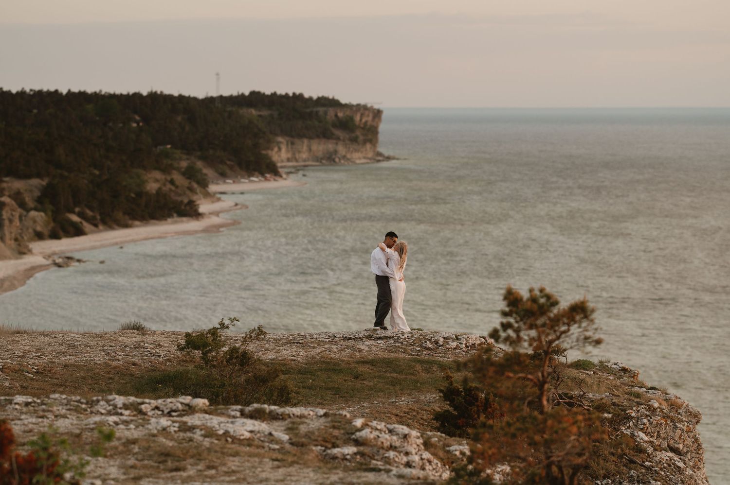 Couple embracing on rocky coastal cliff with turquoise sea and forested shoreline in background.