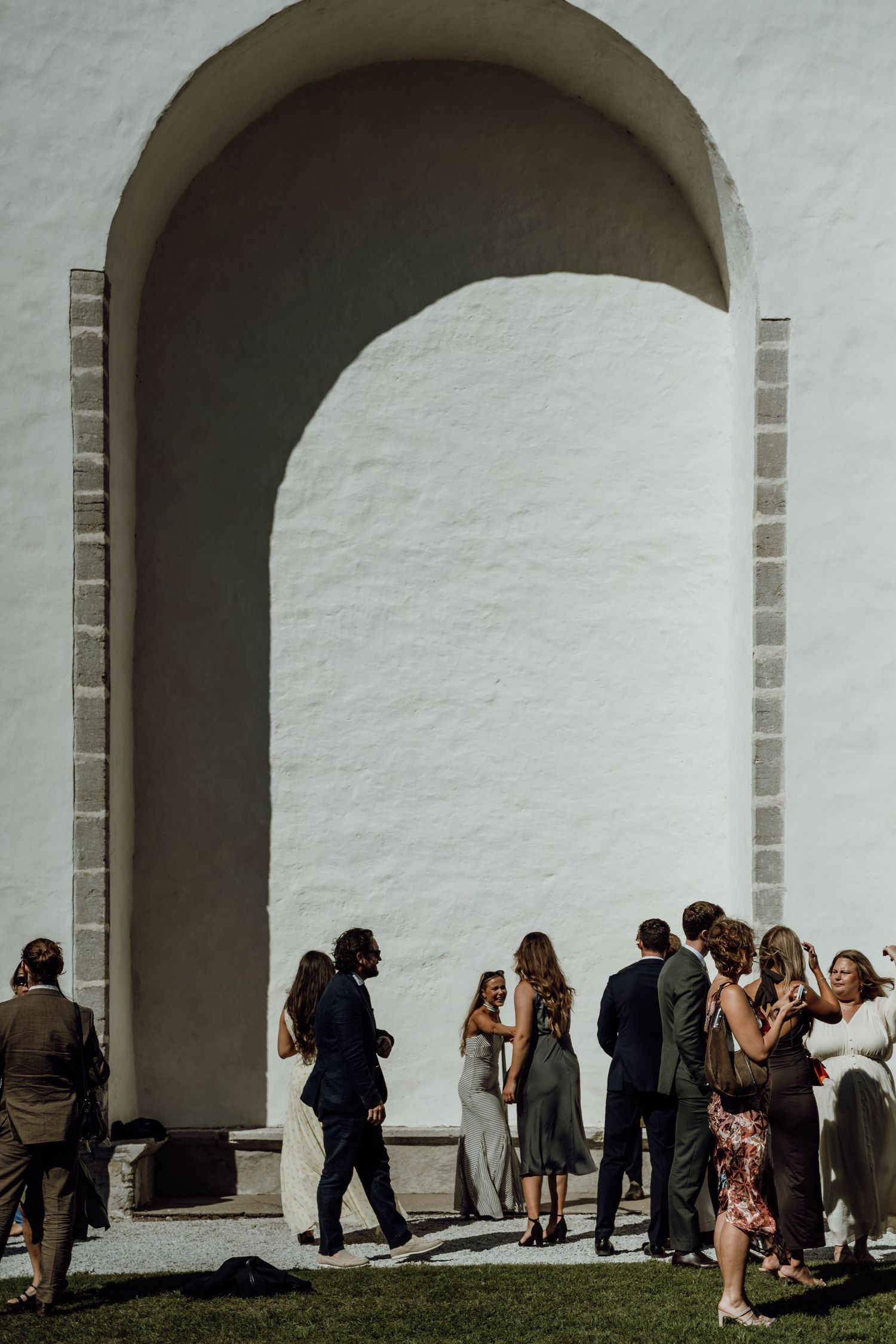 People gather in front of white arched walls during a sunny day, casting dramatic shadows on the architecture.