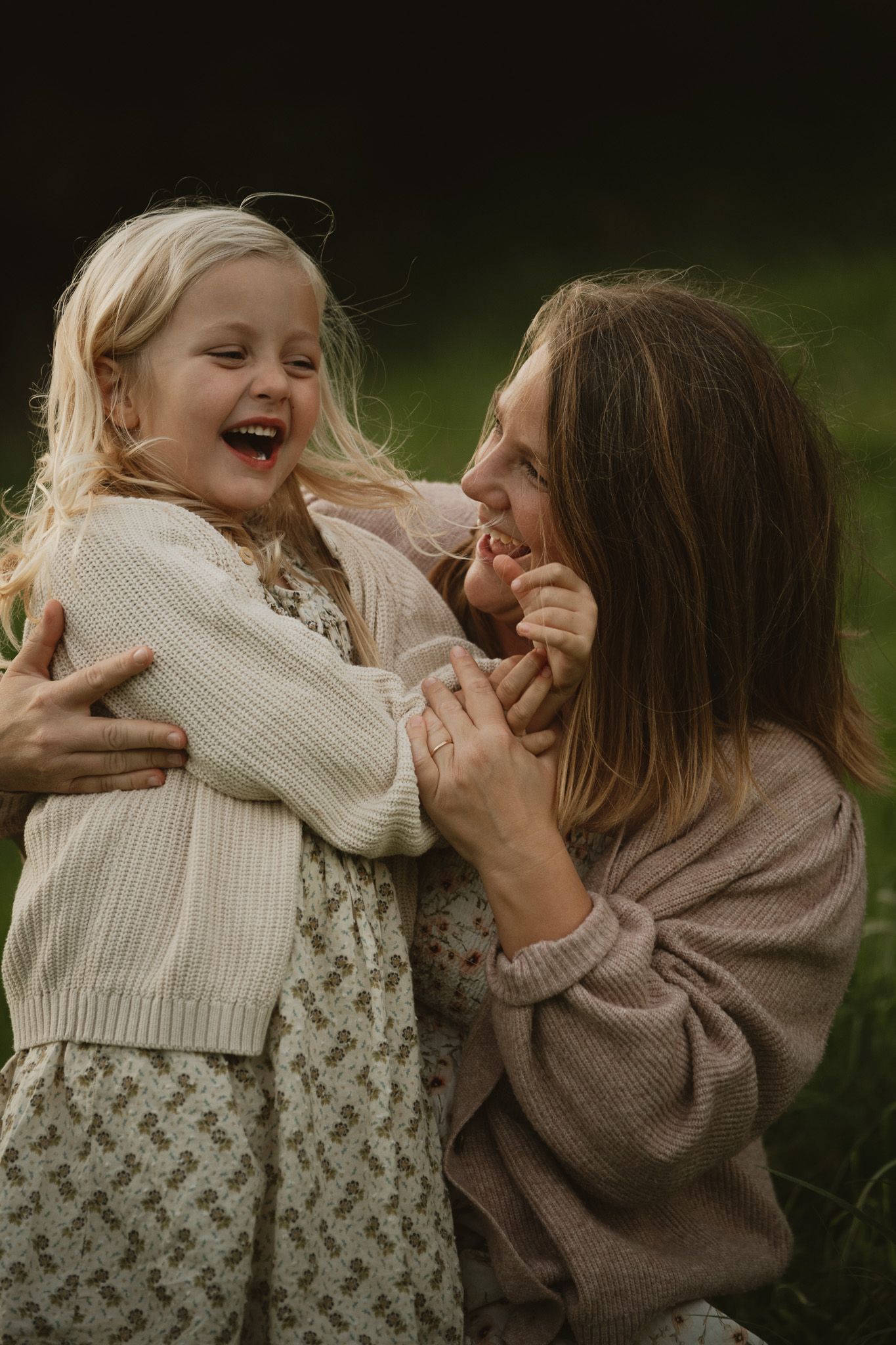 A mother and young blonde girl laugh together outdoors, sharing a joyful embrace in soft natural light.
