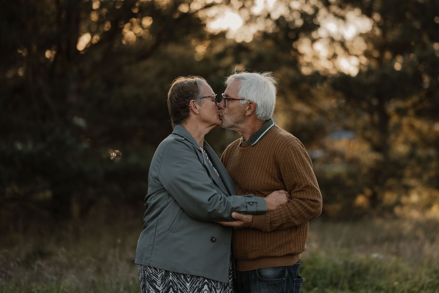 Elderly couple sharing a romantic kiss outdoors, embracing in a golden-hour forest setting.