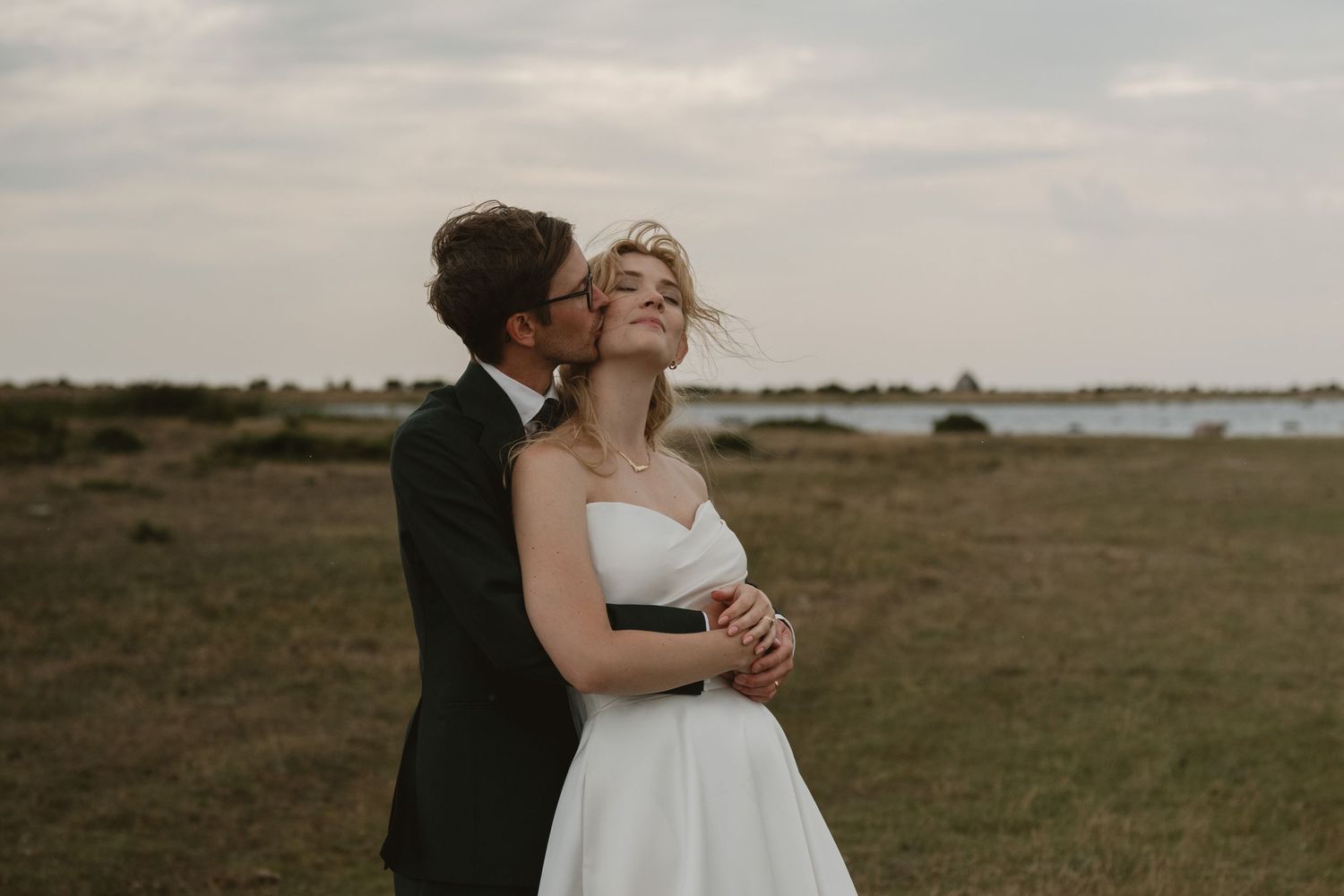 Groom in dark suit kissing bride in white strapless gown outdoors in a scenic field near water.