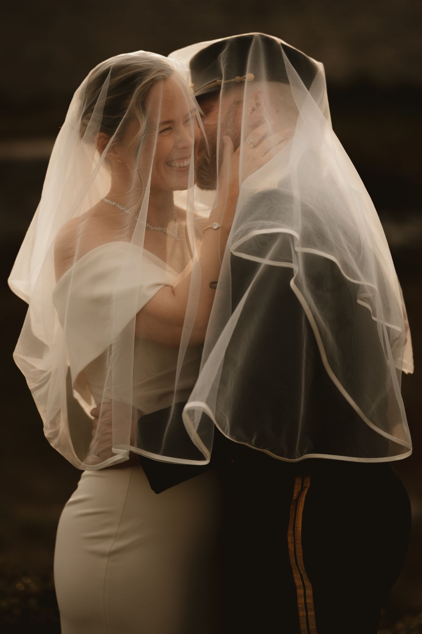 Bride and groom sharing an intimate moment under a flowing white wedding veil, smiling closely together.