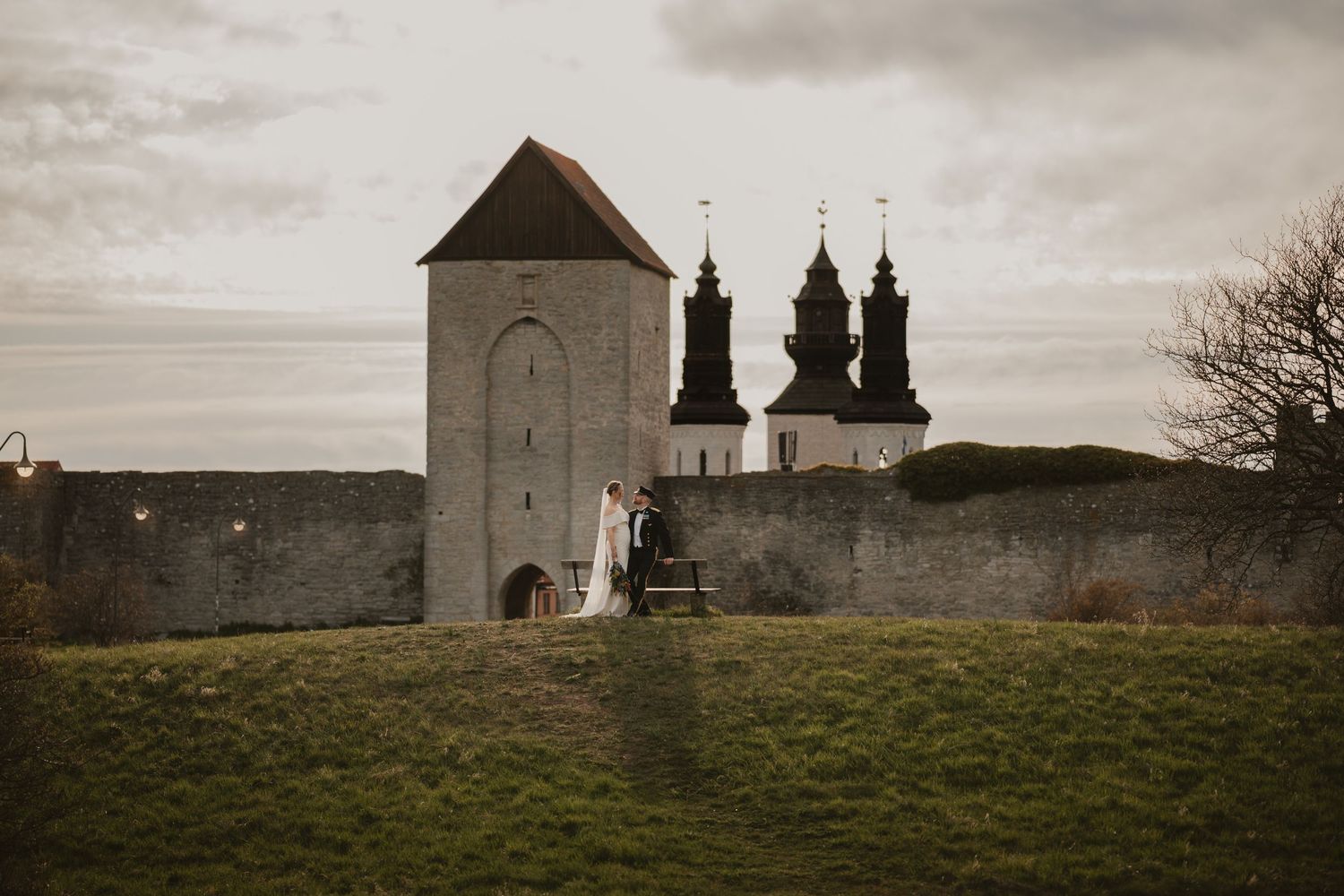 A wedding couple stands on steps beside a medieval stone tower and church in a lush green landscape.