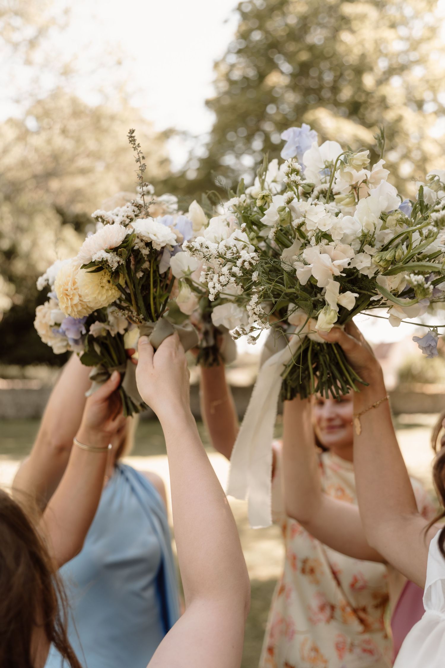 Bridesmaids holding up white floral bouquets against a sunlit outdoor backdrop.