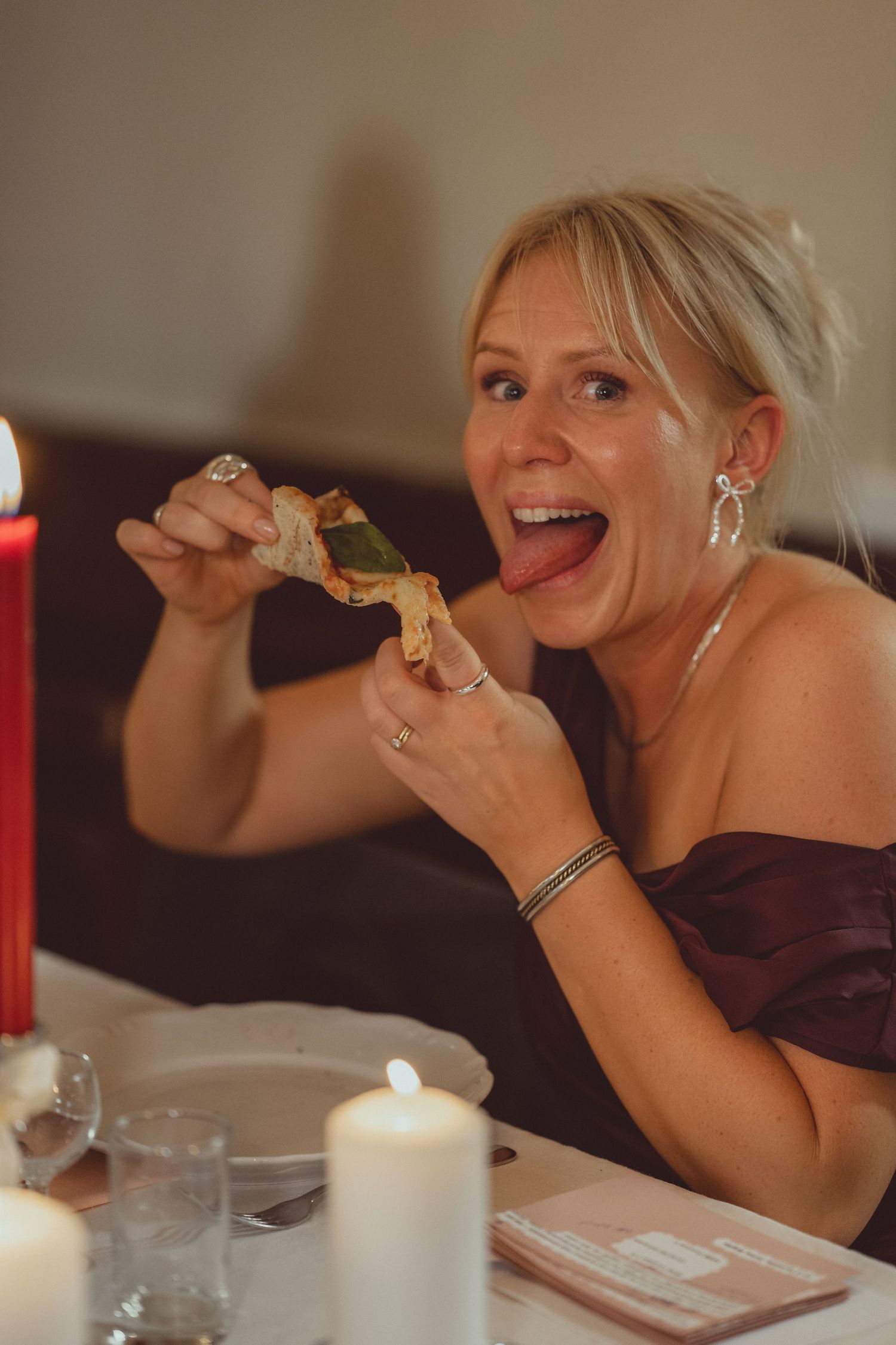 Blonde woman in off-shoulder dress excitedly eating food by candlelight at an elegant dinner table.