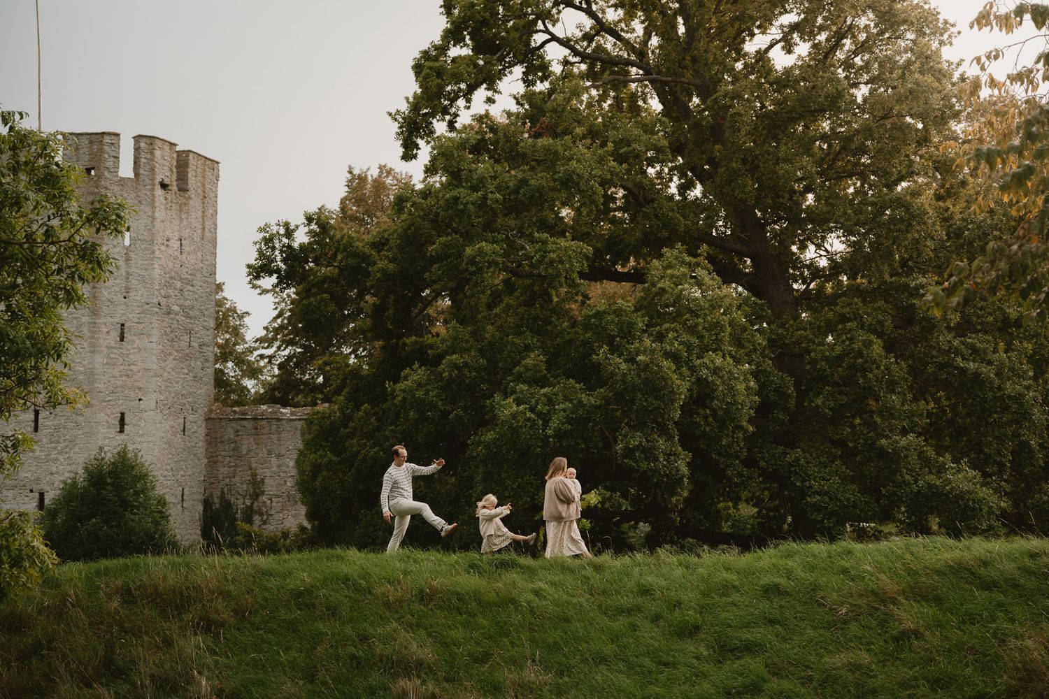 Family playing on green hillside near ancient stone castle surrounded by lush trees at dusk.
