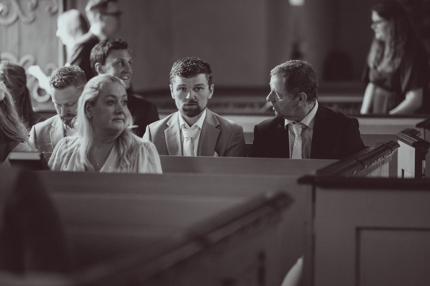 Black and white photo of guests seated in church pews during a wedding ceremony, engaged in quiet conversation.