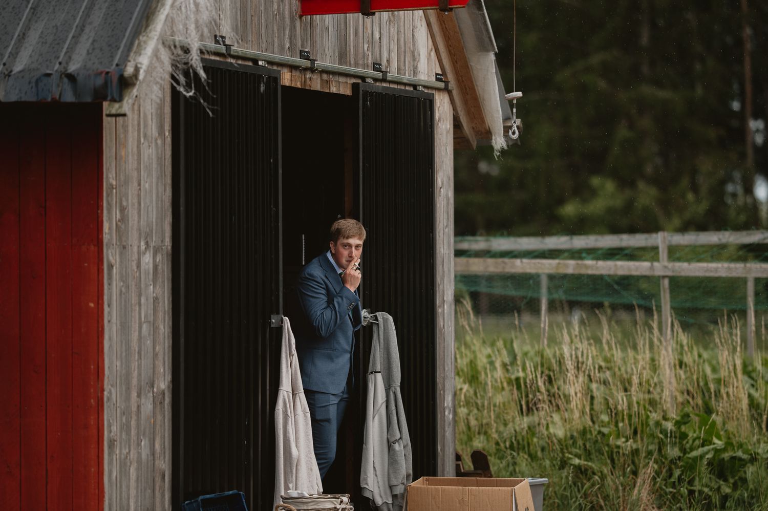 A man in a blue suit talks on phone in a rustic barn doorway surrounded by lush green grass and red wooden walls.