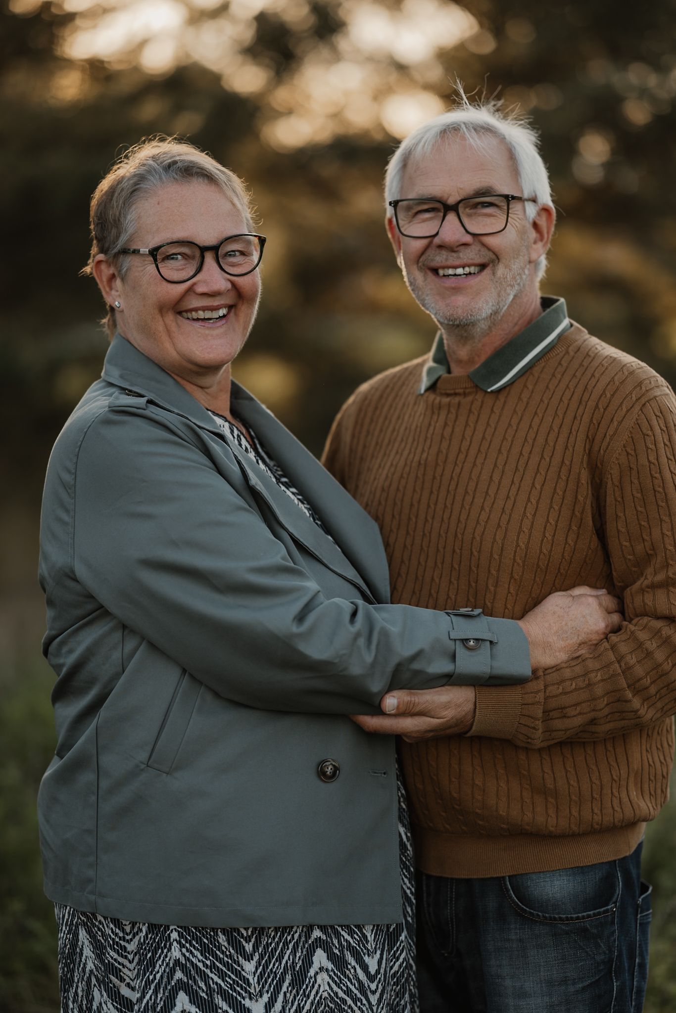 Smiling older couple with glasses embracing outdoors in warm golden light, wearing casual autumn attire.