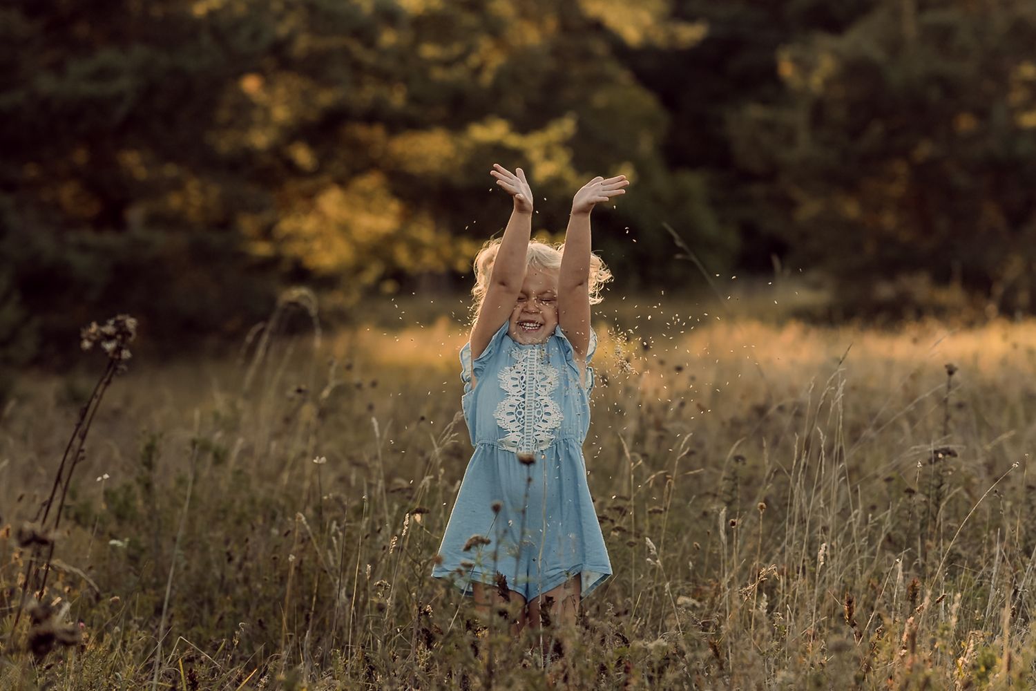 A joyful young girl in a blue dress plays in a sunlit meadow with arms raised, surrounded by tall grass and trees.