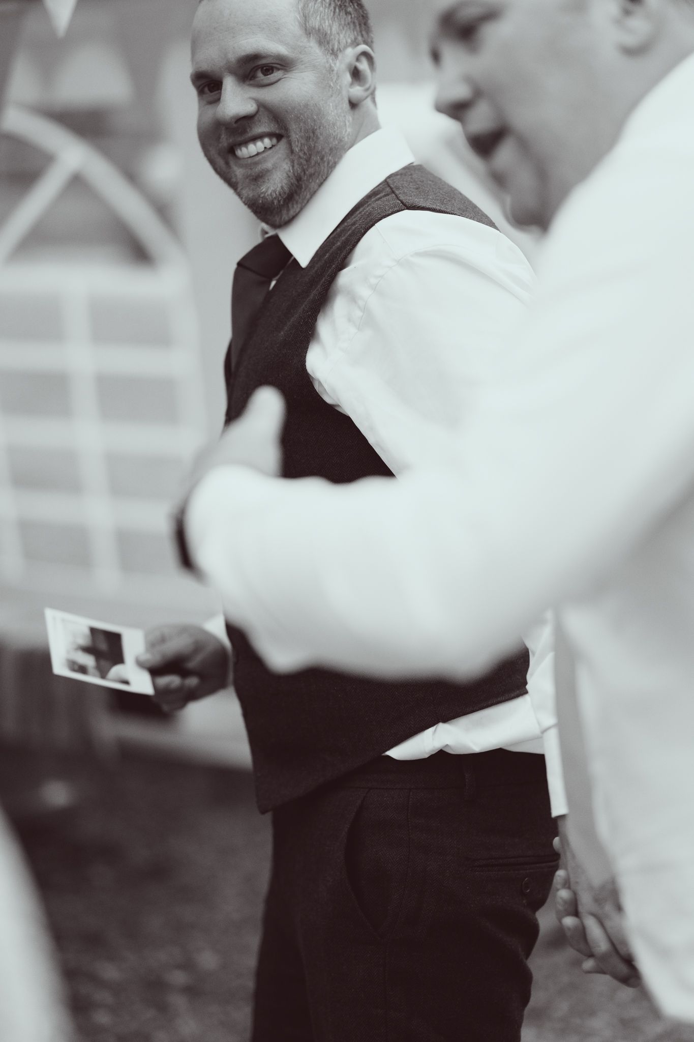 Black and white photo of a smiling bearded groom in vest and tie holding a card at a wedding.