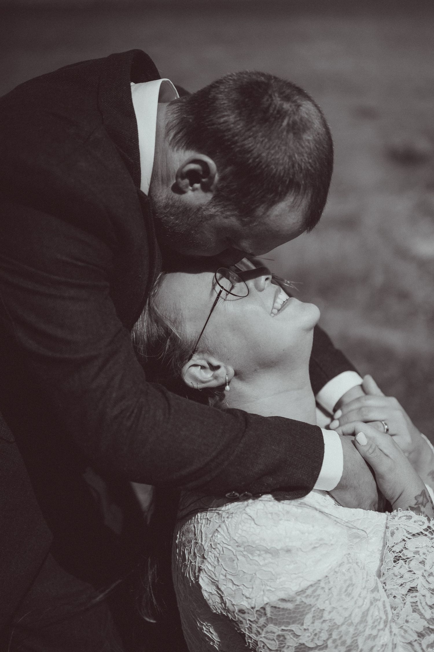 Black and white wedding photo of groom leaning in to kiss smiling bride in lace dress.