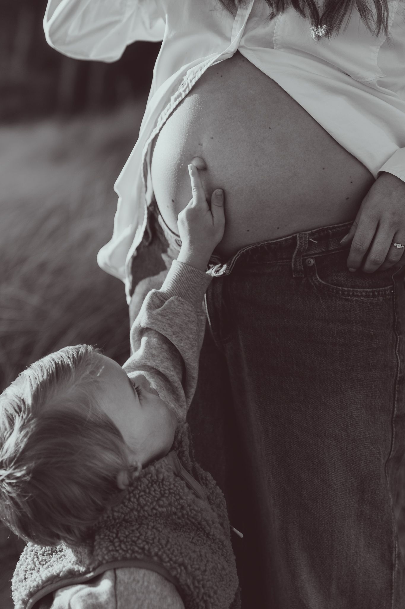 Black and white photo of a child touching a pregnant woman's bare belly outdoors.