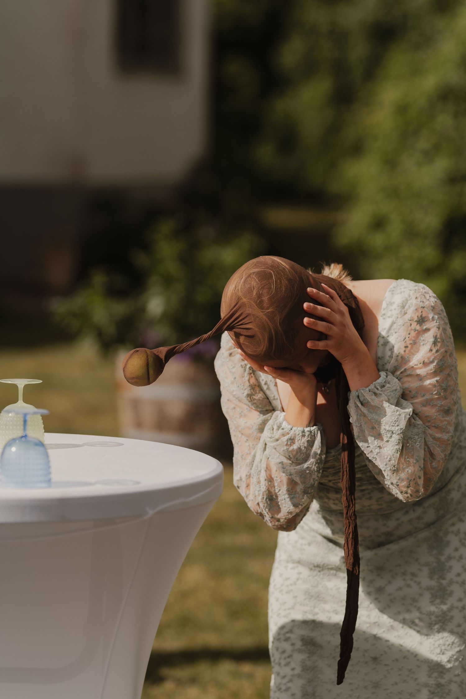 Woman in floral dress bending over outdoor table with hand sanitizer dispenser in sunny garden setting.