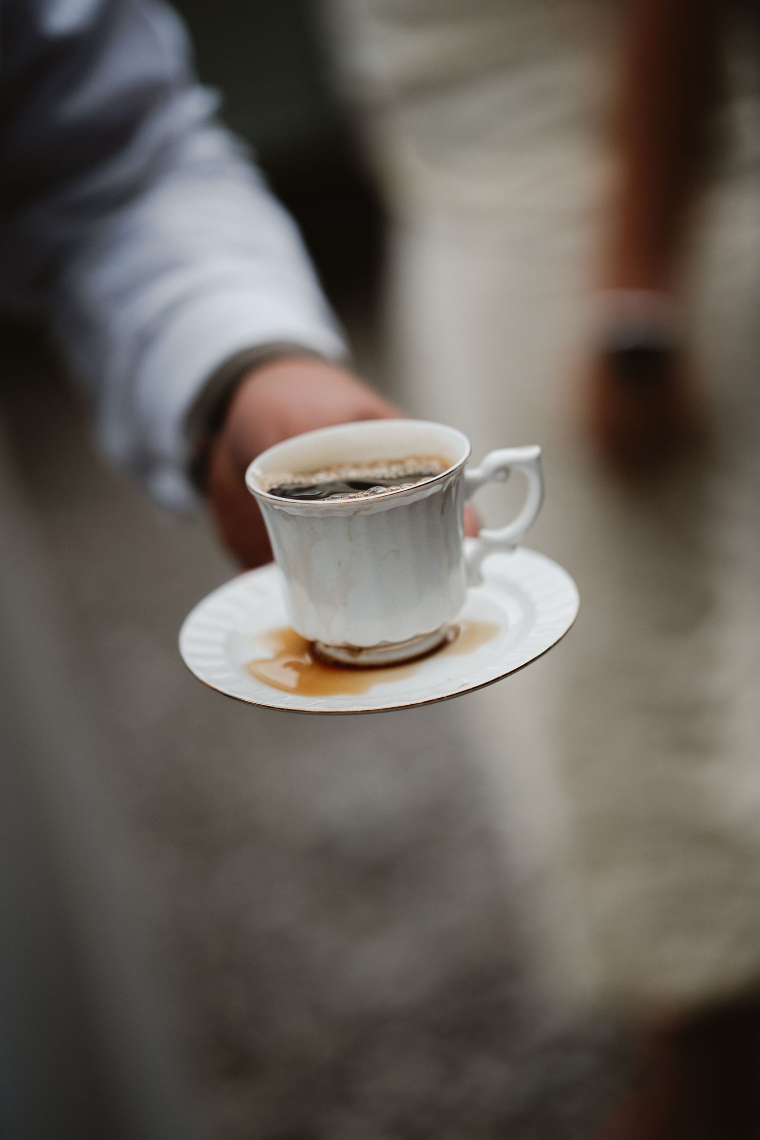 A hand holding a white porcelain cup and saucer with black coffee, showing a small spill on the saucer.