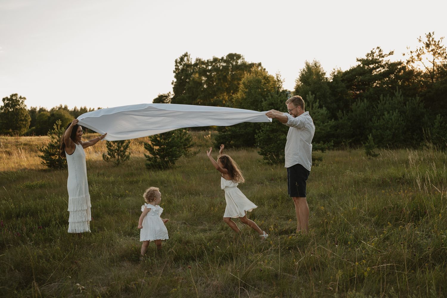 Family playing with white fabric in a sunny meadow at golden hour, parents and two young girls in white dresses.
