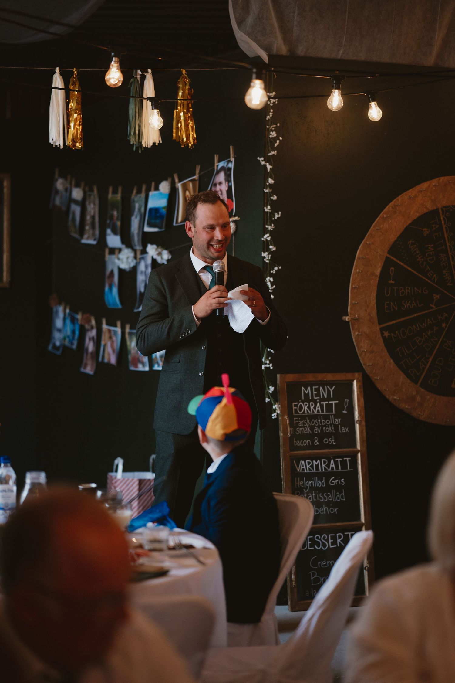 A man in a suit gives a speech at a wedding reception with string lights and a chalkboard menu in the background.
