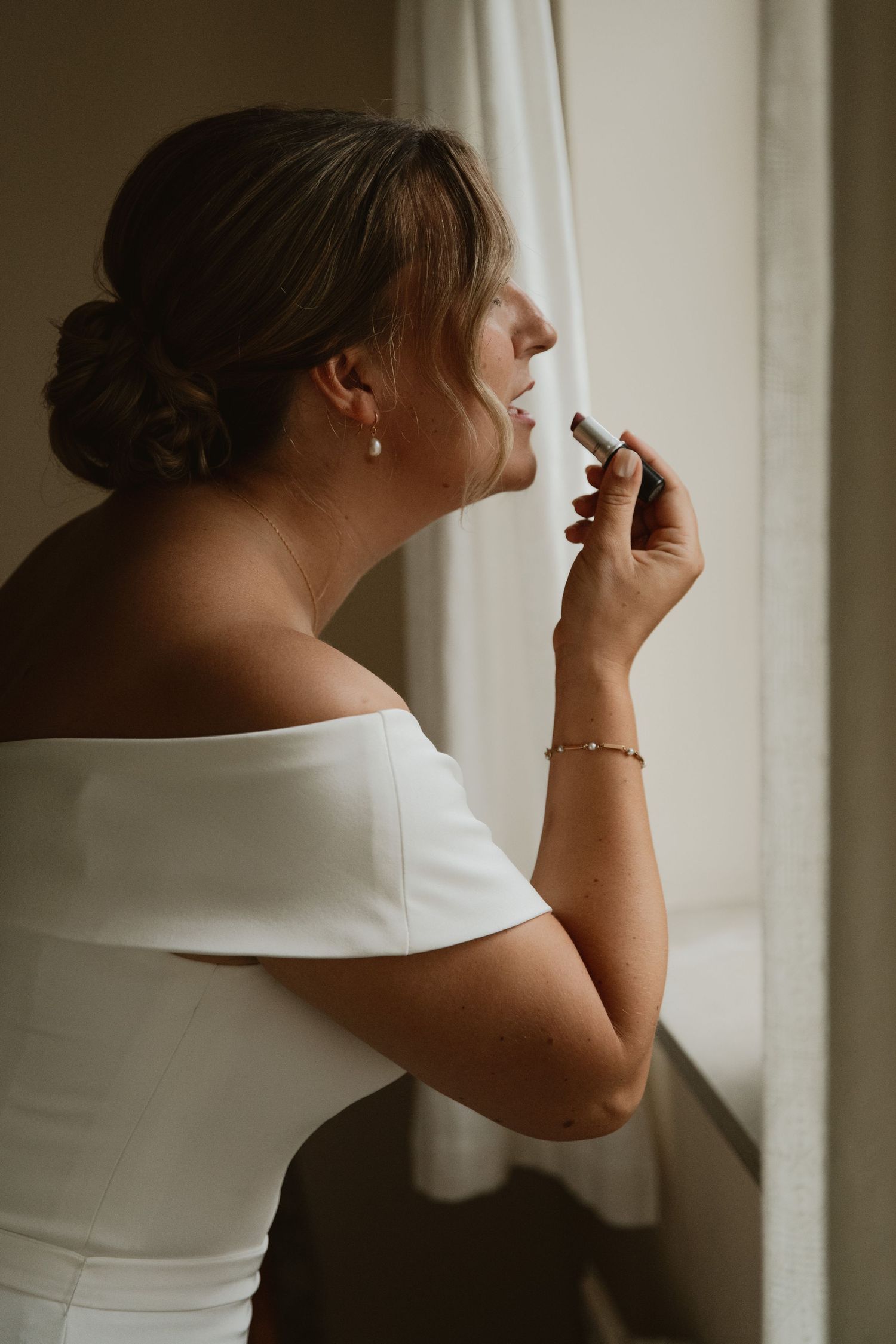 Bride in white off-shoulder gown applying lipstick by a window on her wedding day.