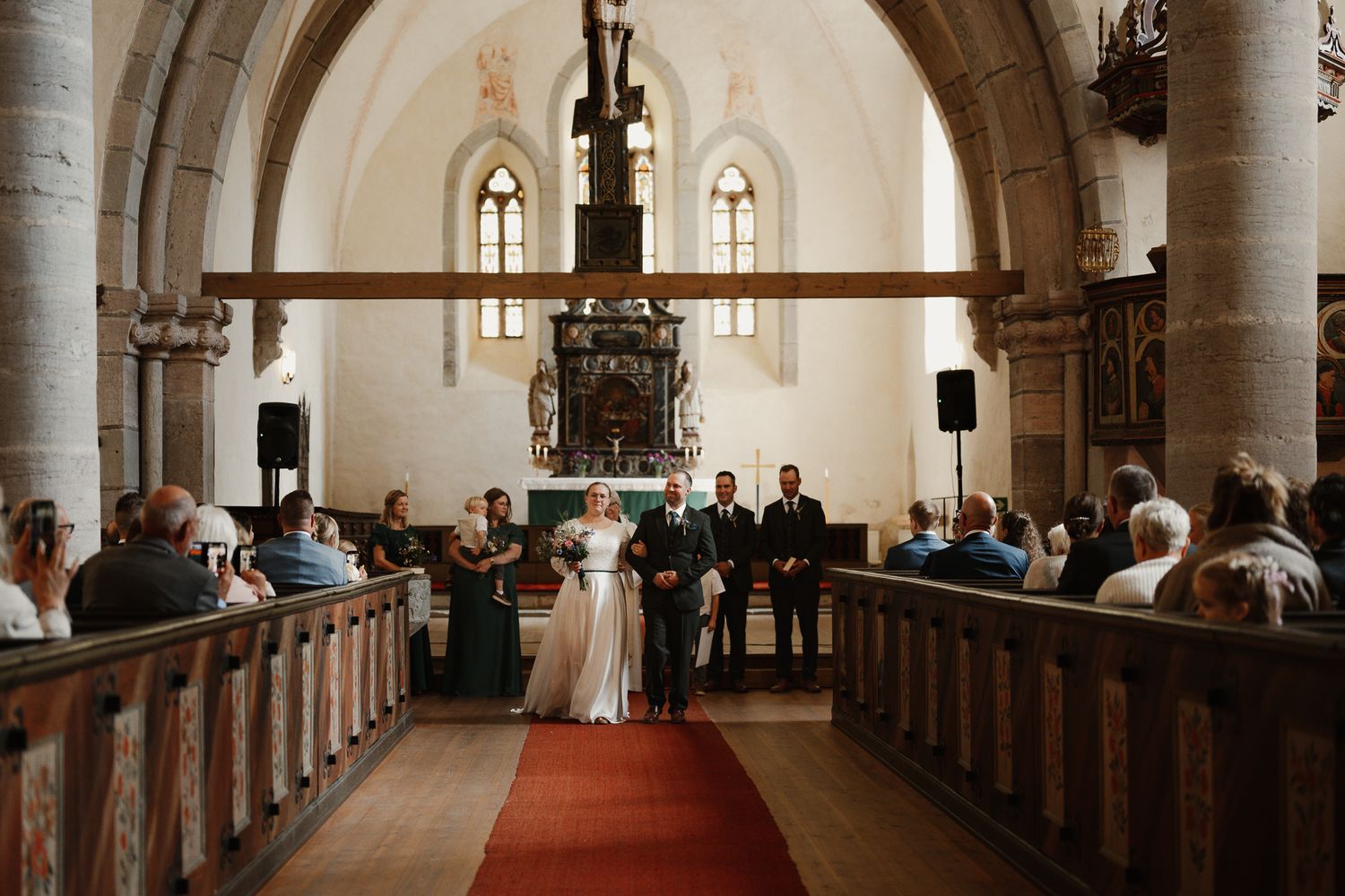 Bride and groom stand at the altar of a historic stone church with red carpet aisle and wooden pews filled with guests.