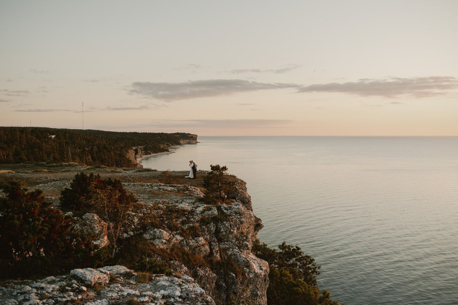A couple stands on rocky cliffs overlooking a calm sea at sunset with soft pastel skies in the background.