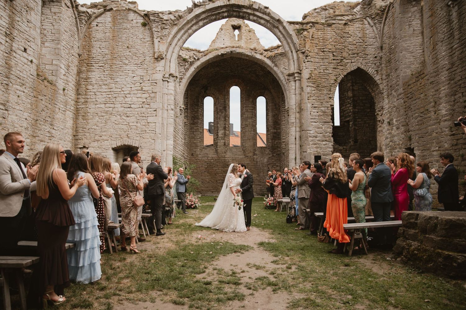 Bride and groom kiss at altar in stunning stone church ruins wedding ceremony with guests applauding.