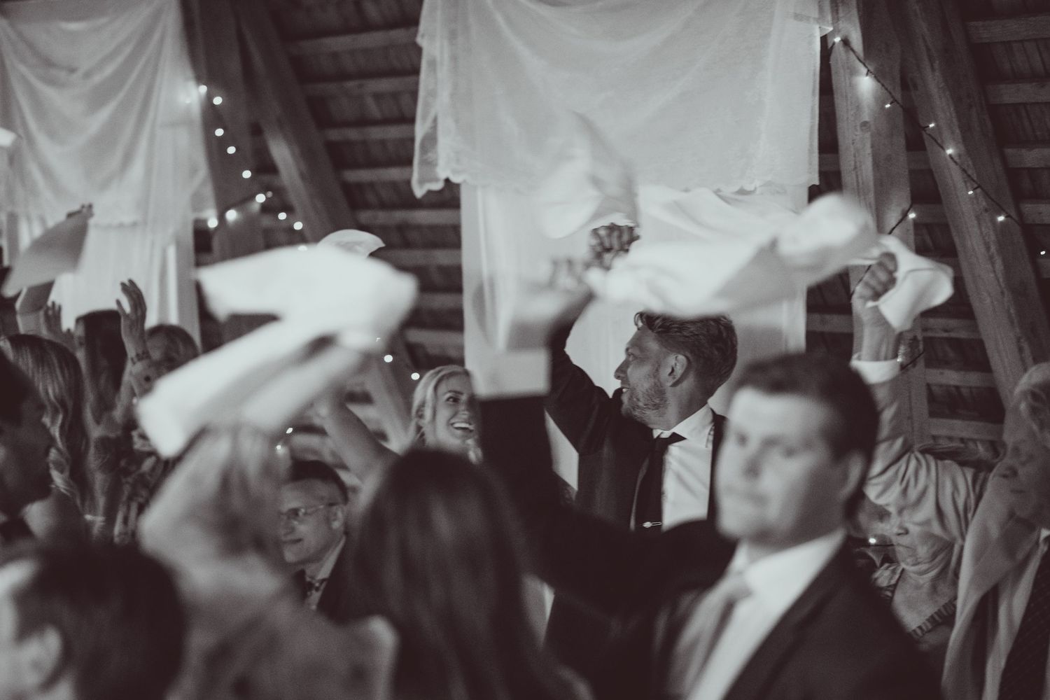 Black and white photo of wedding guests waving white napkins on the dance floor at a rustic barn reception.