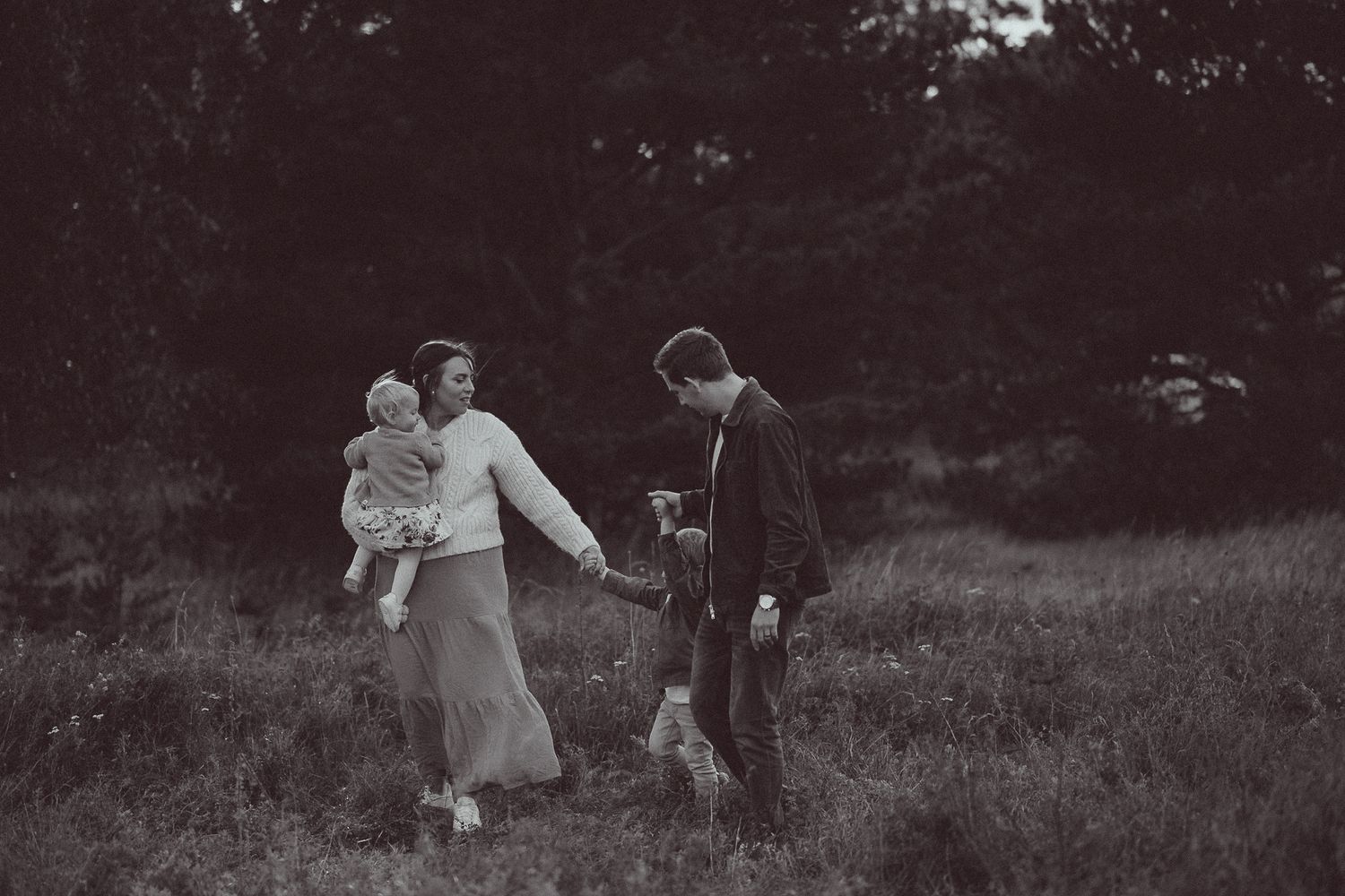 Black and white family photo of parents and two young children walking through a grassy field at dusk.