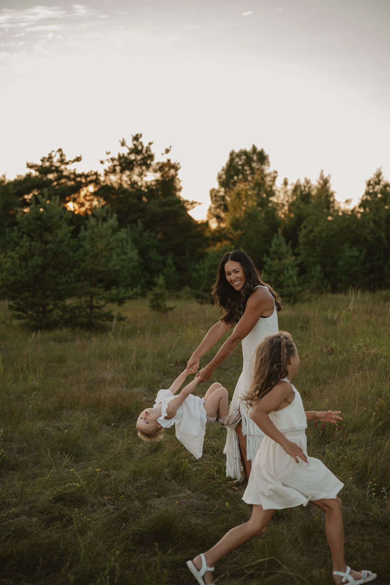 A mother joyfully swings her toddler while her daughter runs nearby in a sunlit green meadow at sunset.