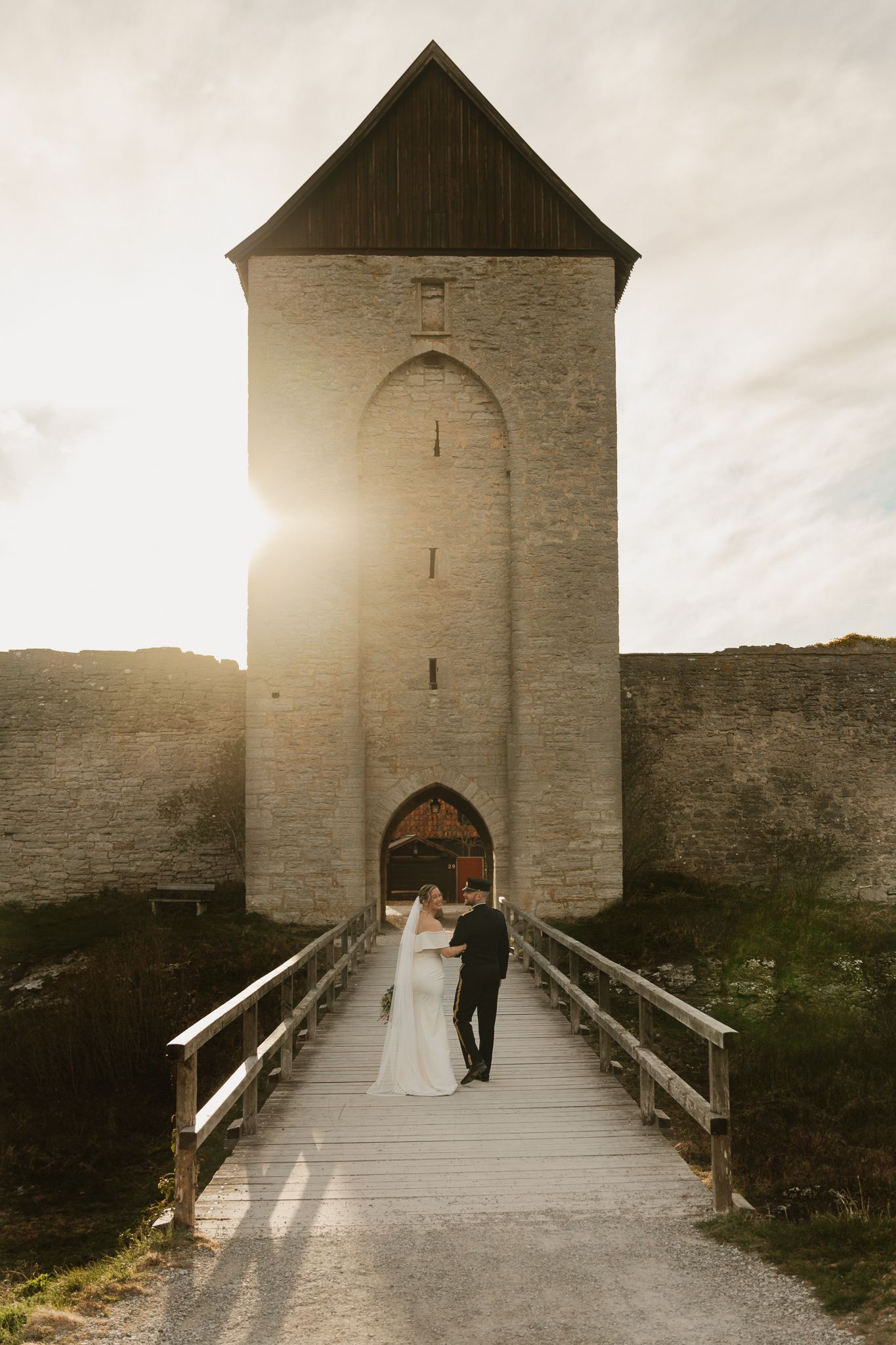 Bride and groom stand on a wooden bridge before a sunlit medieval stone tower at golden hour.