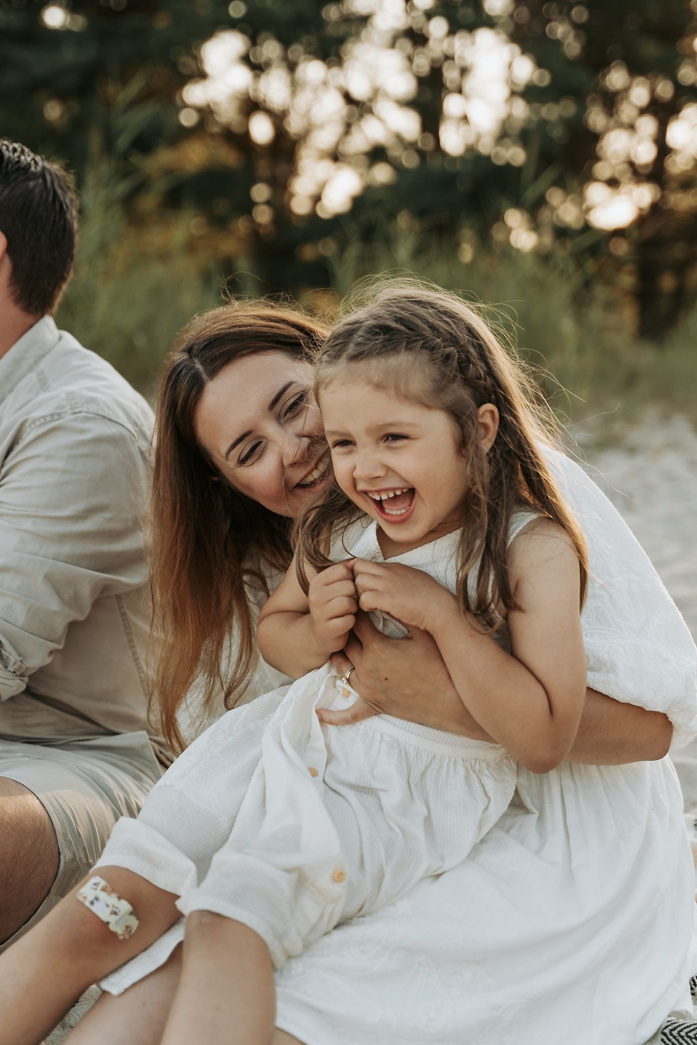 A joyful mother hugs her laughing little girl in a white dress on a sandy beach surrounded by trees.