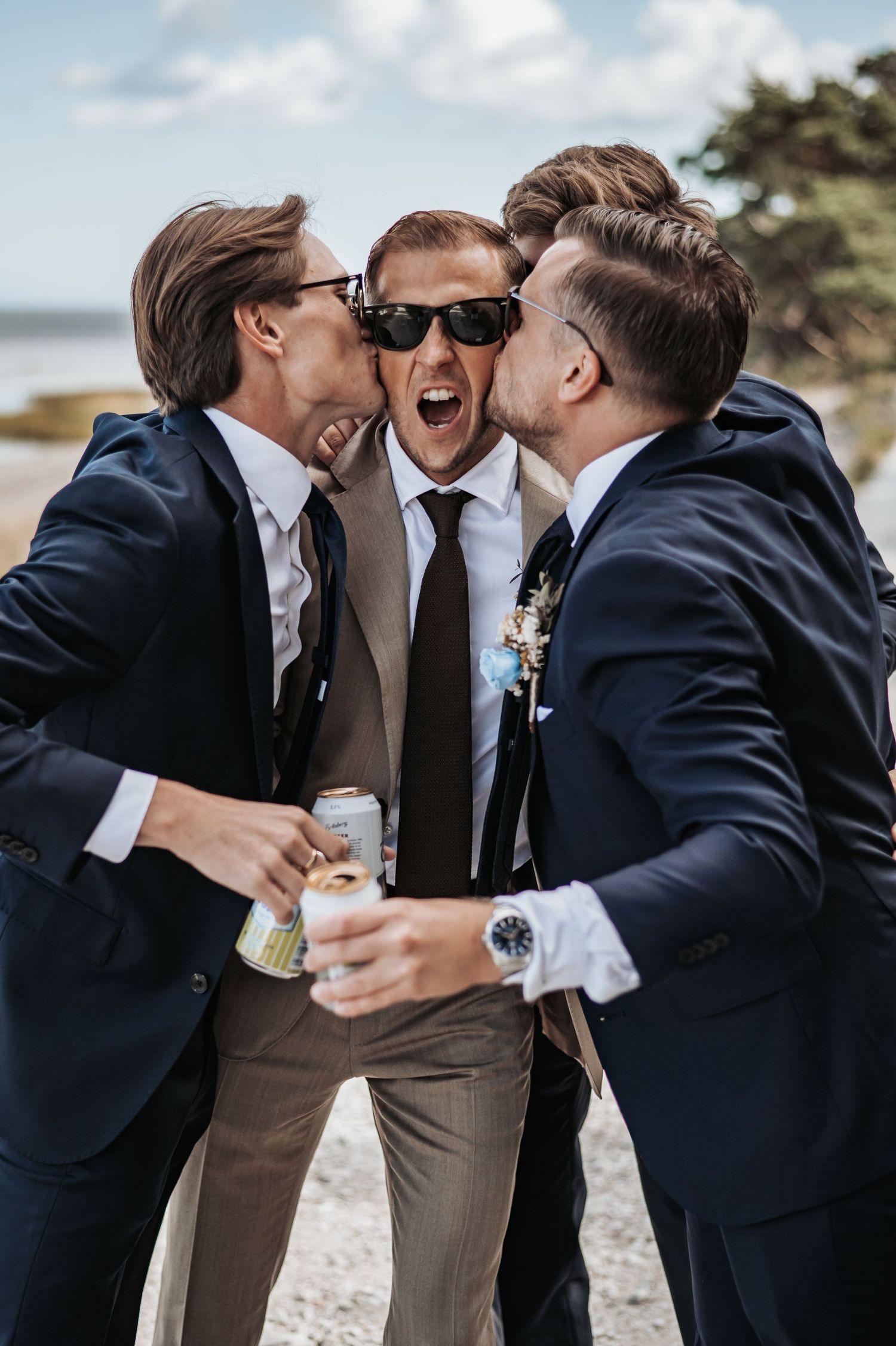 Groomsmen in navy suits sharing laughs and drinks on a beach.