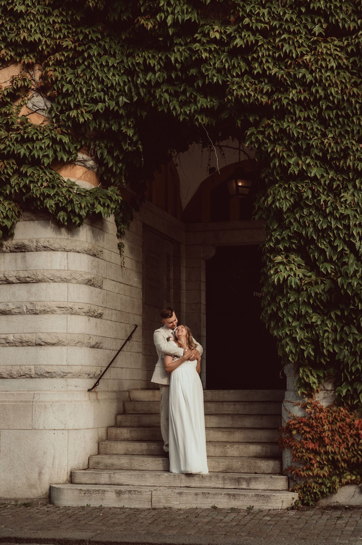 Romantic wedding couple embraces in a stone archway surrounded by lush green ivy and foliage.