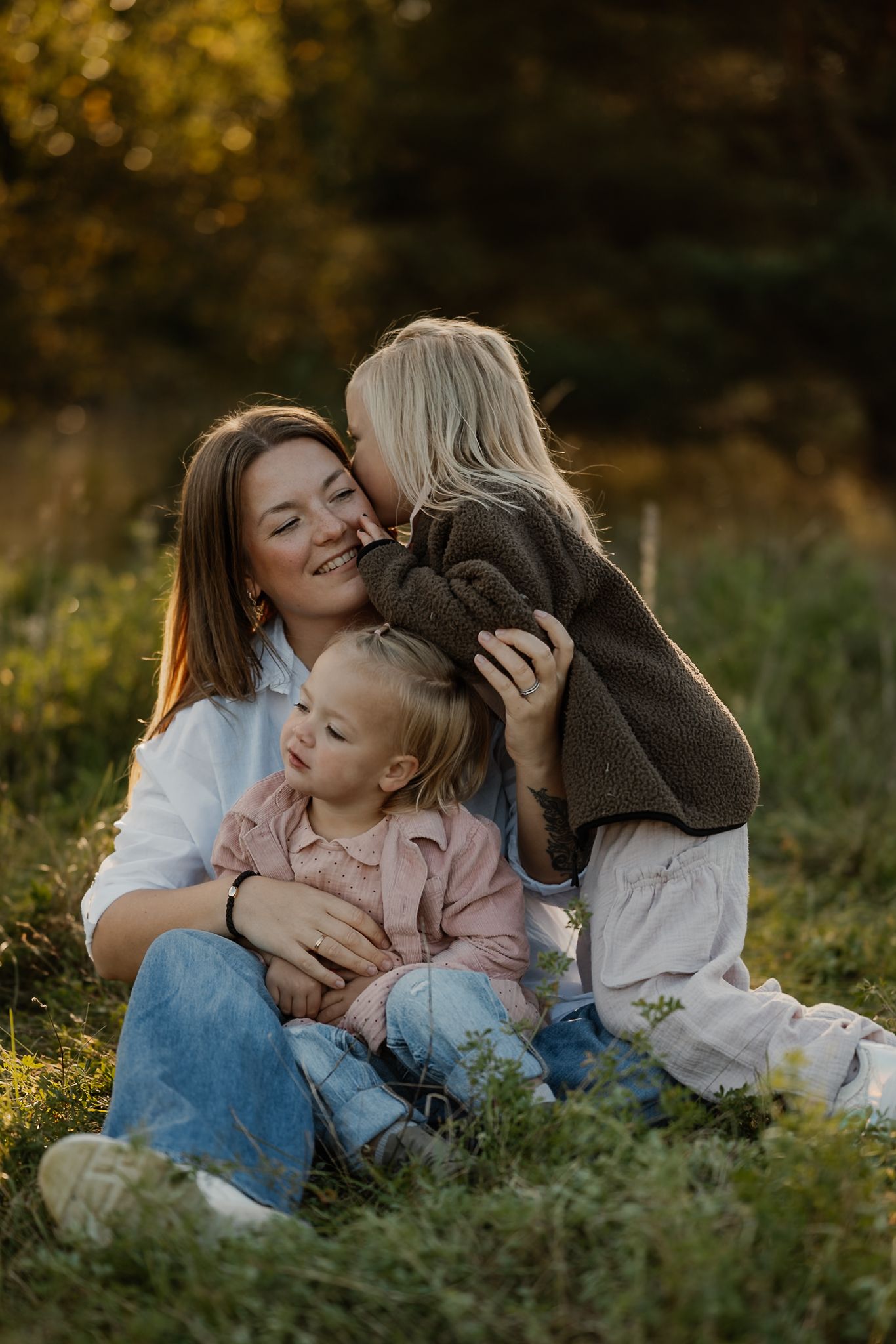 A smiling mother sits in a sunny meadow hugging two young blonde children, one kissing her cheek.