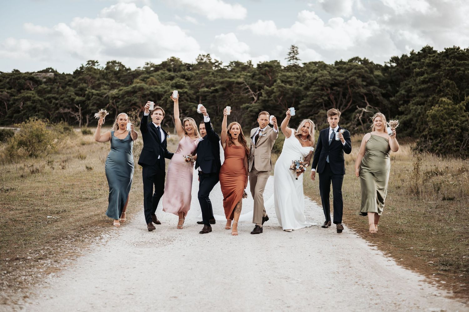 Wedding party celebrates joyfully on a dirt path surrounded by green trees and nature.