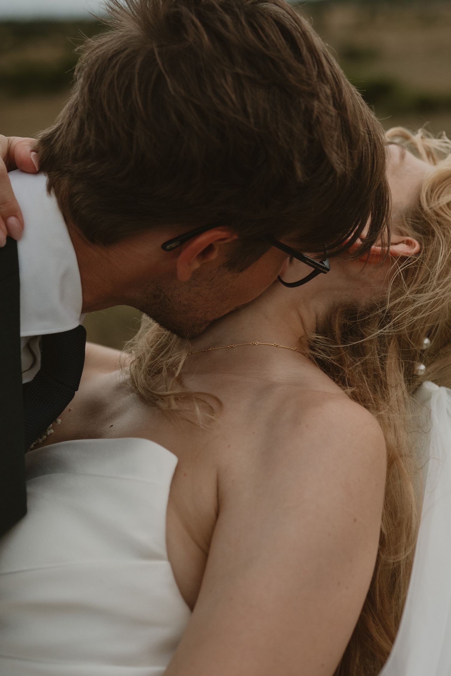 Groom in glasses kissing bride on neck during romantic outdoor wedding portrait session.