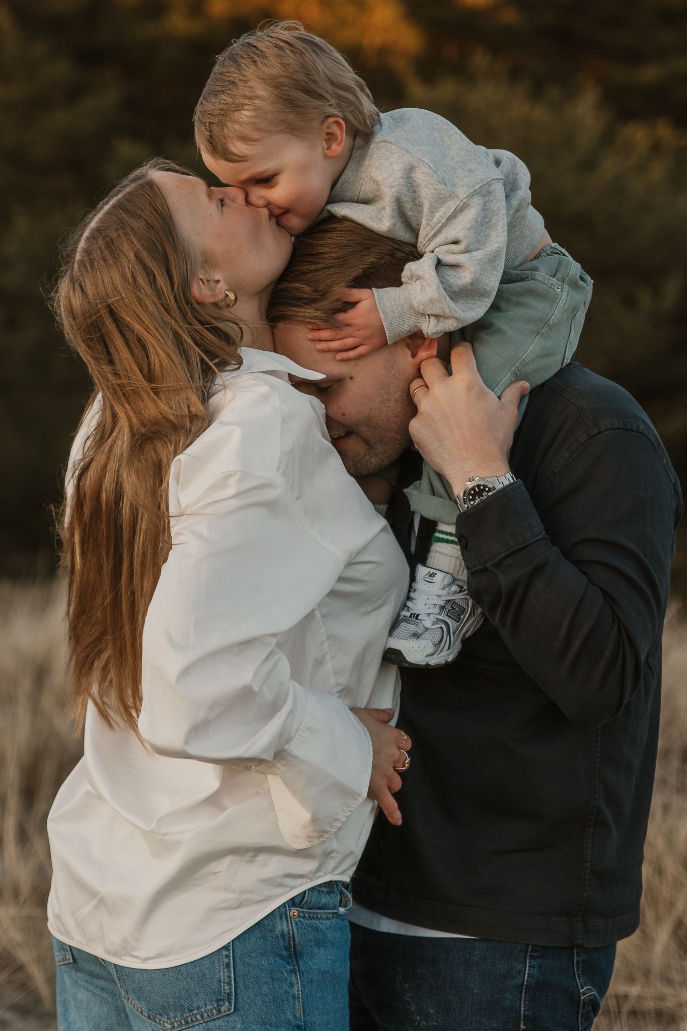A loving family shares a sweet moment outdoors, with parents kissing their young child in a warm autumn setting.