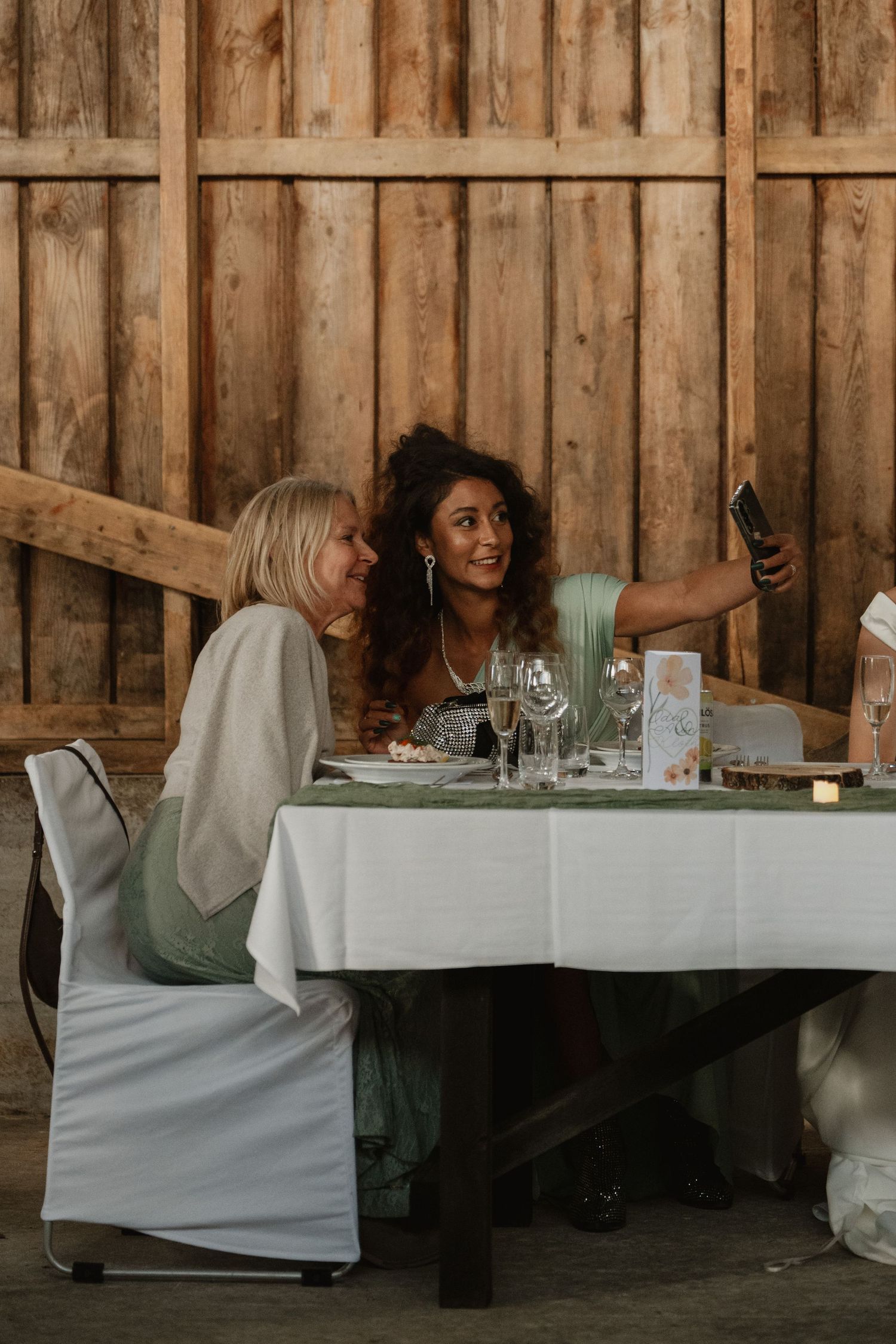 Two women taking a selfie at a decorated dining table inside a rustic wooden barn venue.