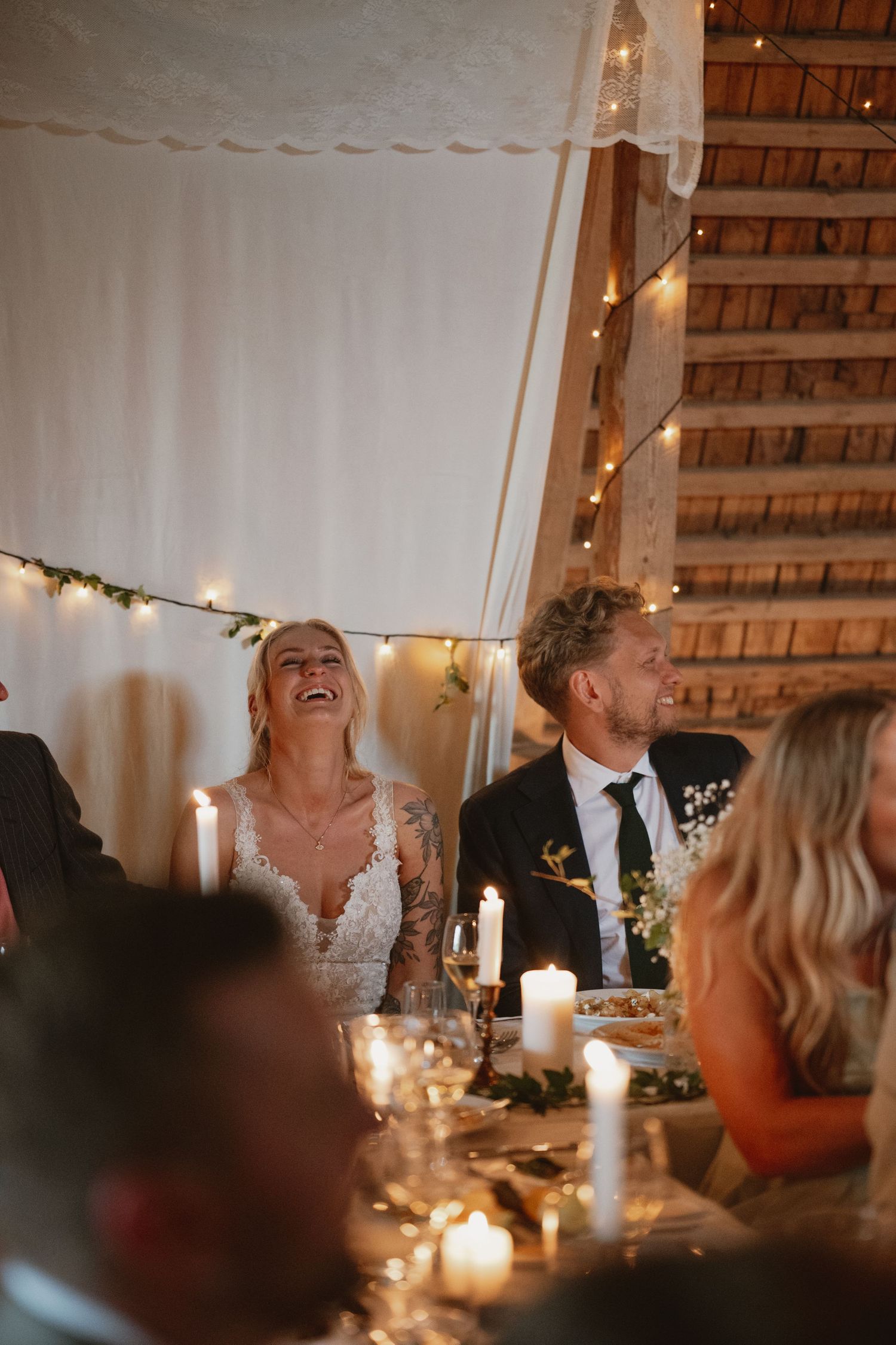 Bride laughing joyfully with groom at candlelit wedding reception in rustic barn venue with fairy lights.
