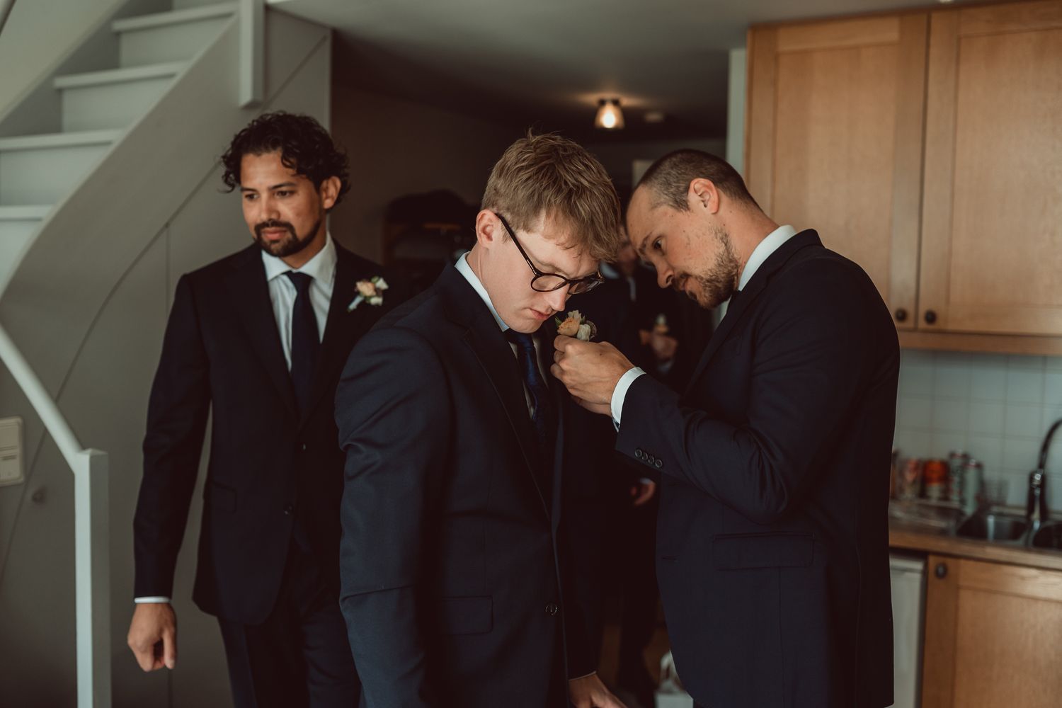A group gets ready in a home kitchen wearing formal black suits.