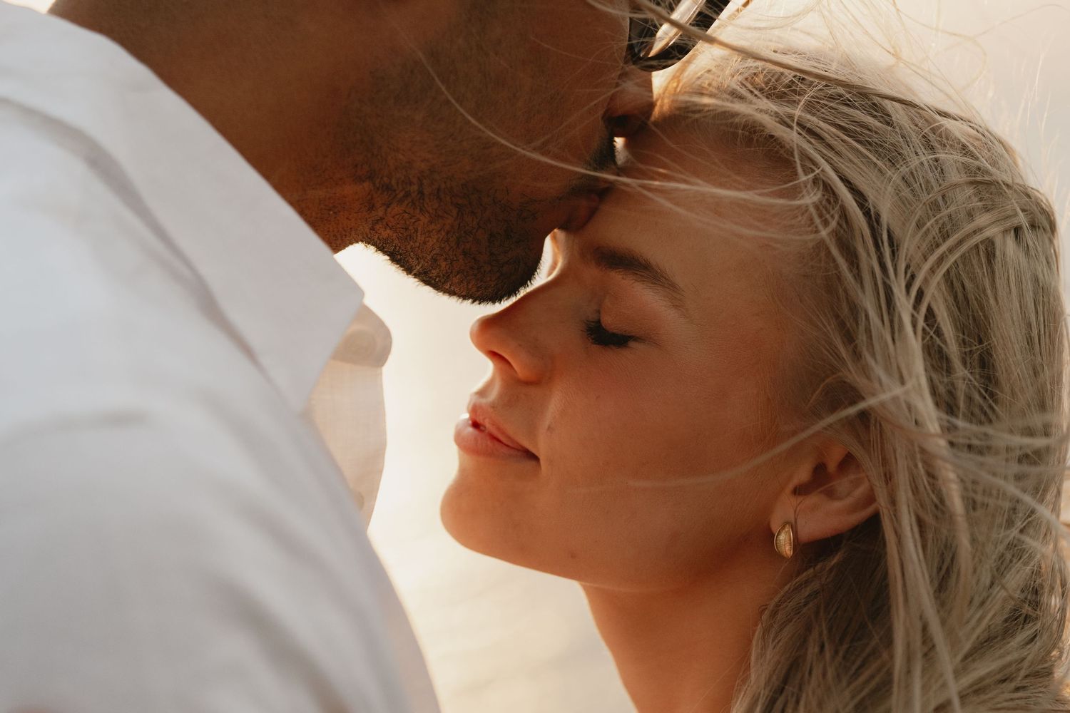 Romantic couple sharing an intimate moment, nearly kissing, with soft natural lighting and windswept blonde hair.