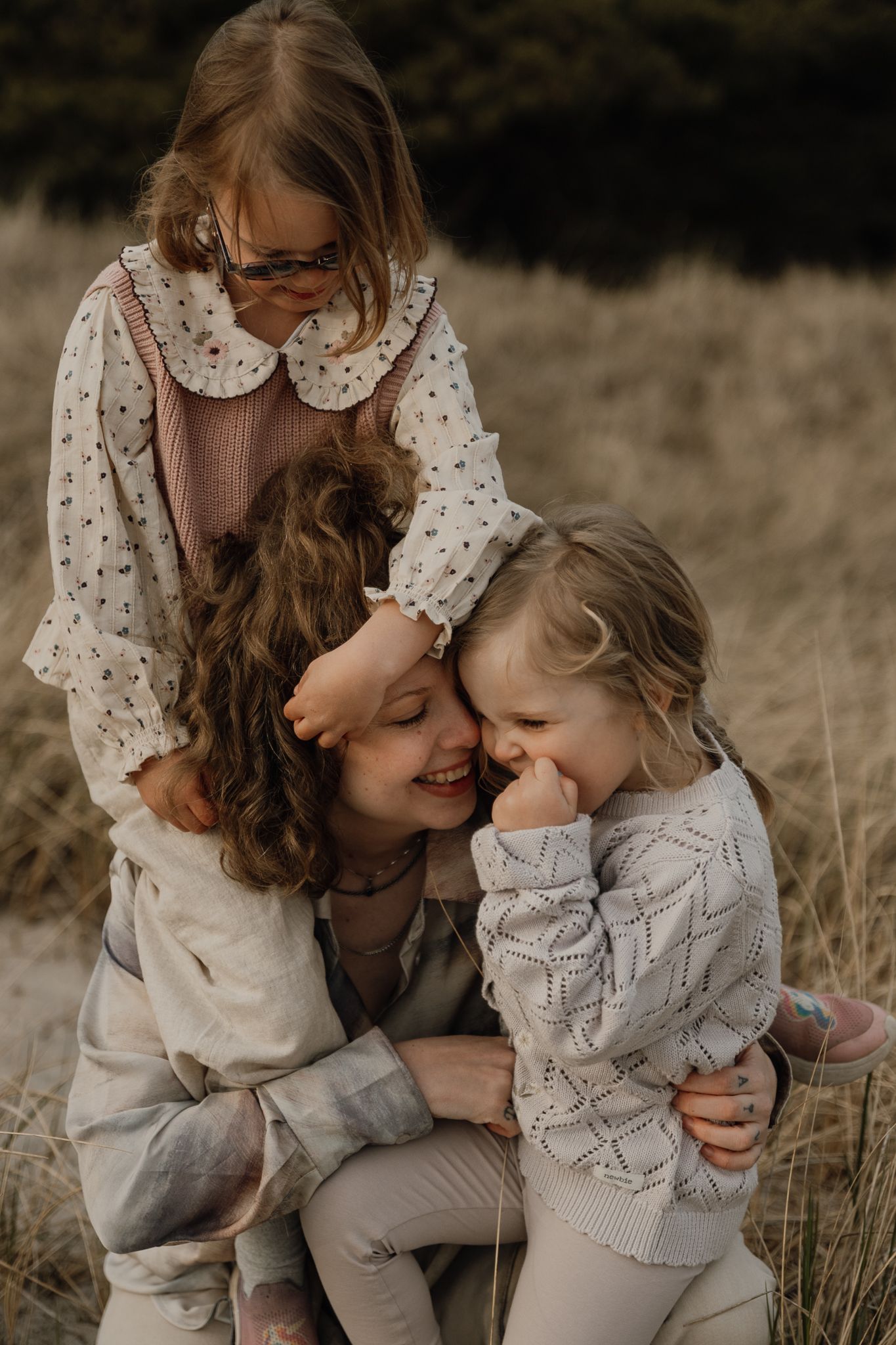 A mother crouching in a field embracing her two young daughters, sharing a joyful moment outdoors.