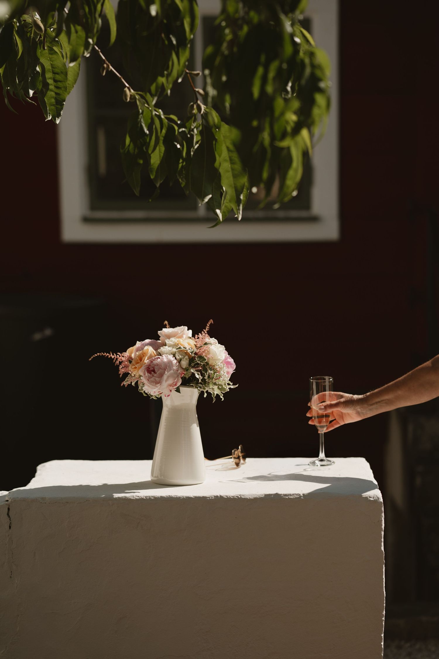 A white pitcher with pink floral arrangement on a draped table outdoors, with a hand holding a champagne flute nearby.
