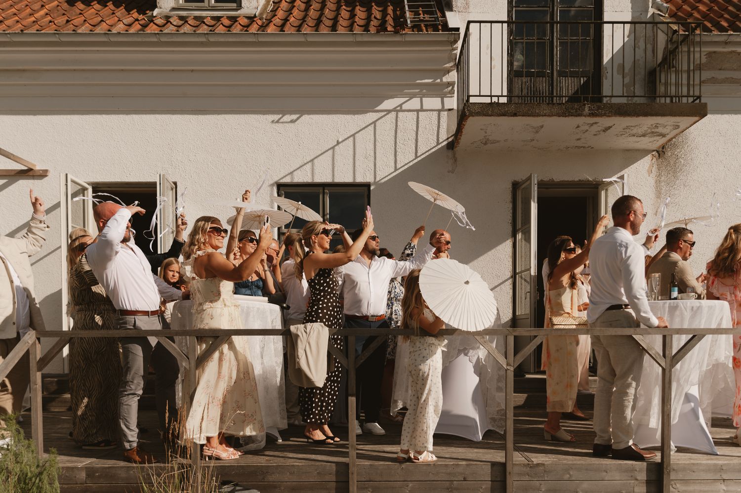 Guests in white attire celebrate outdoors on a sunny terrace, raising parasols and glasses at an elegant summer wedding reception.