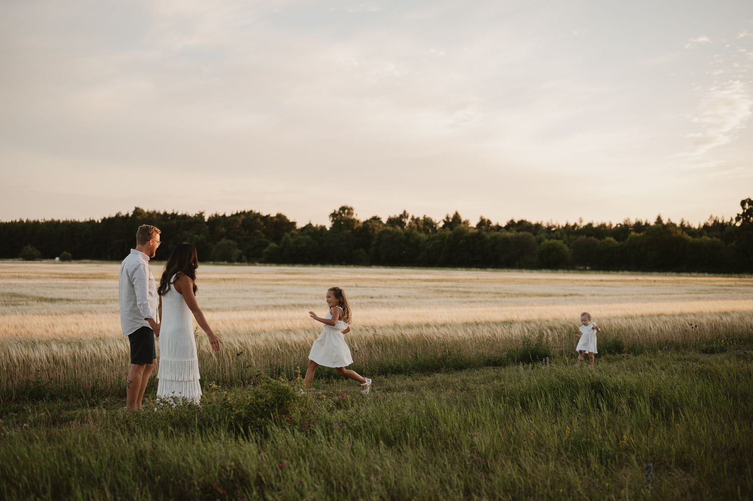 Family of four walking through a lush green field at sunset, parents holding hands with young children in white dresses.
