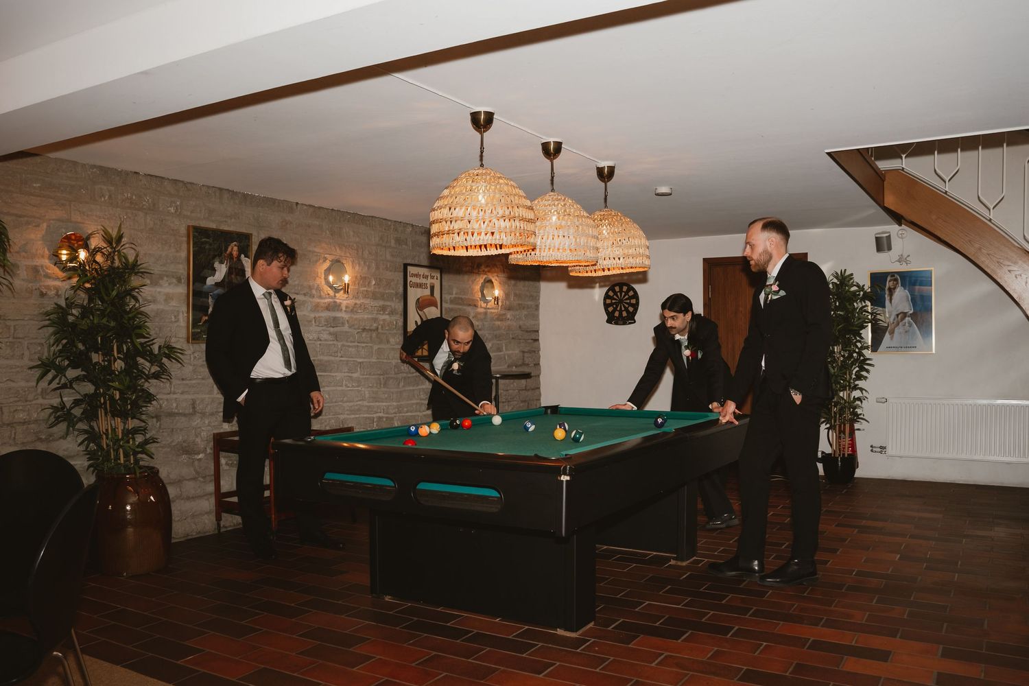Guests in formal attire playing pool on a green billiard table in a stylish venue with wicker pendant lights.
