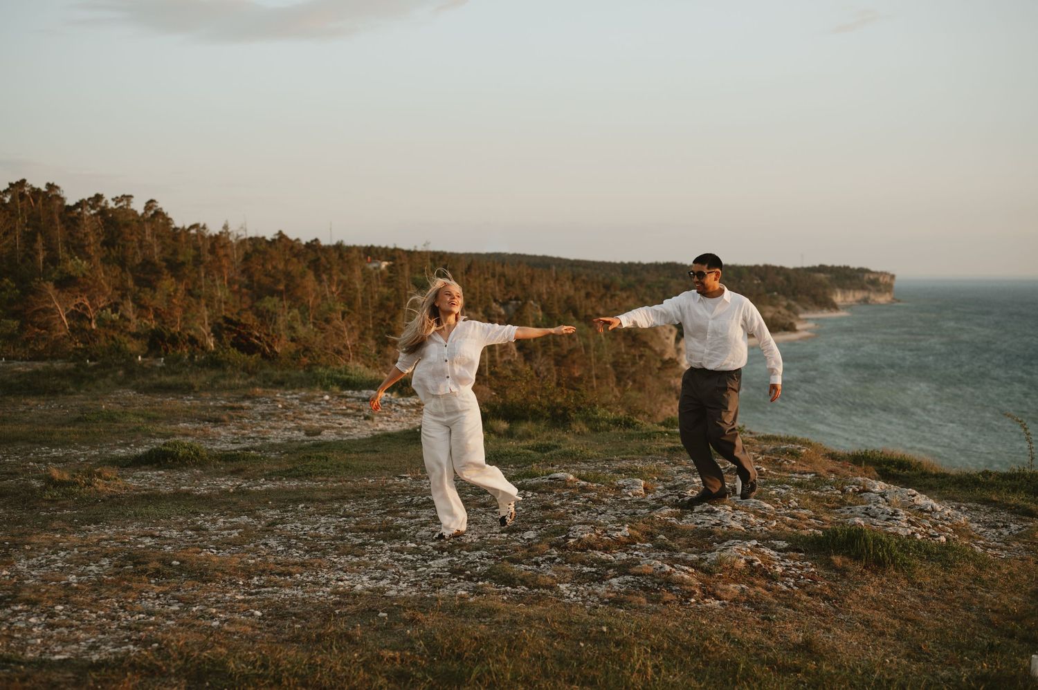 Couple dancing joyfully on rocky coastal cliff with ocean and forest backdrop at golden hour.