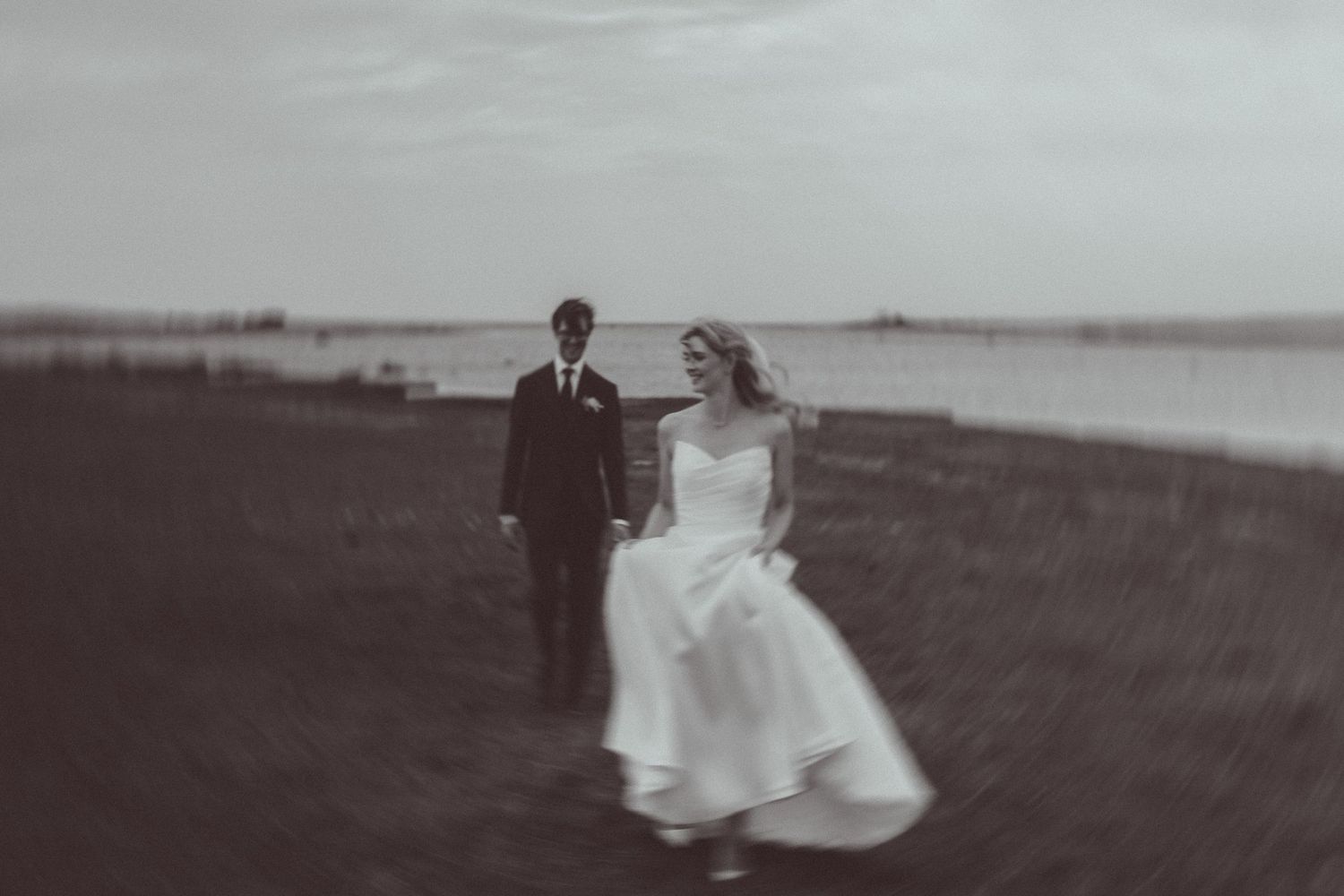 Black and white photo of bride and groom walking near waterfront, wedding dress flowing in the breeze.