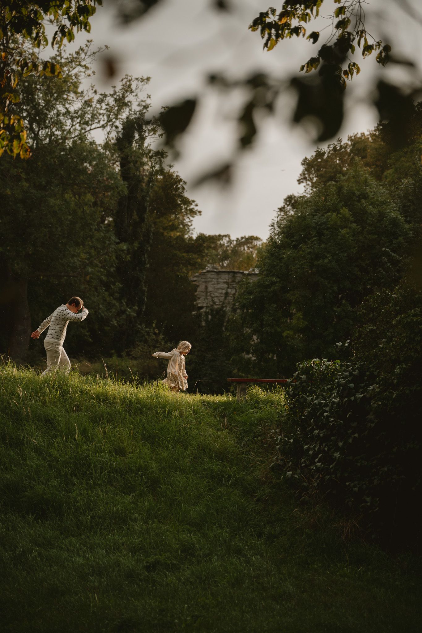 Two people playing on a lush green hillside surrounded by trees in soft evening light.