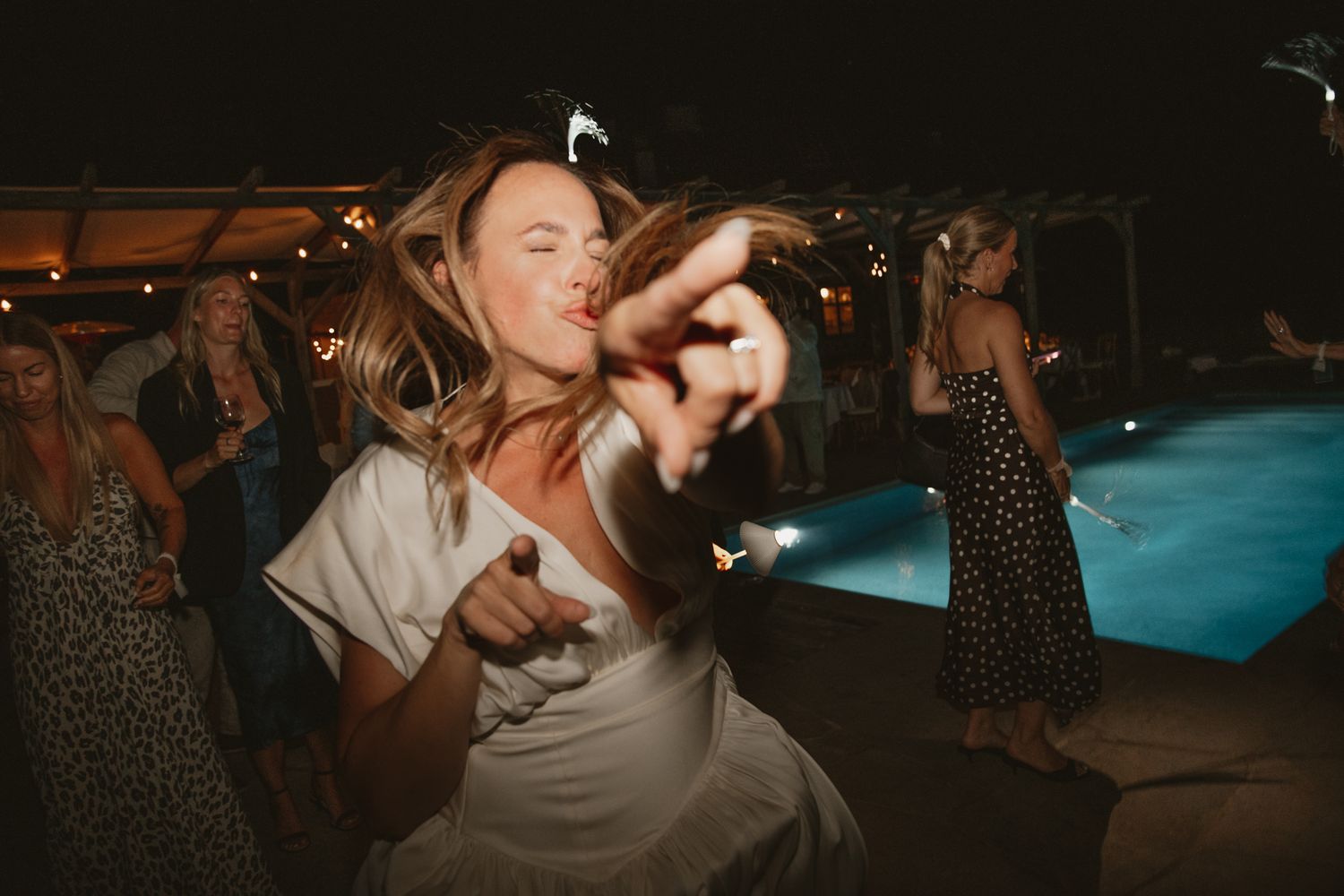 Woman in white dress dancing energetically at an outdoor pool party at night with string lights.